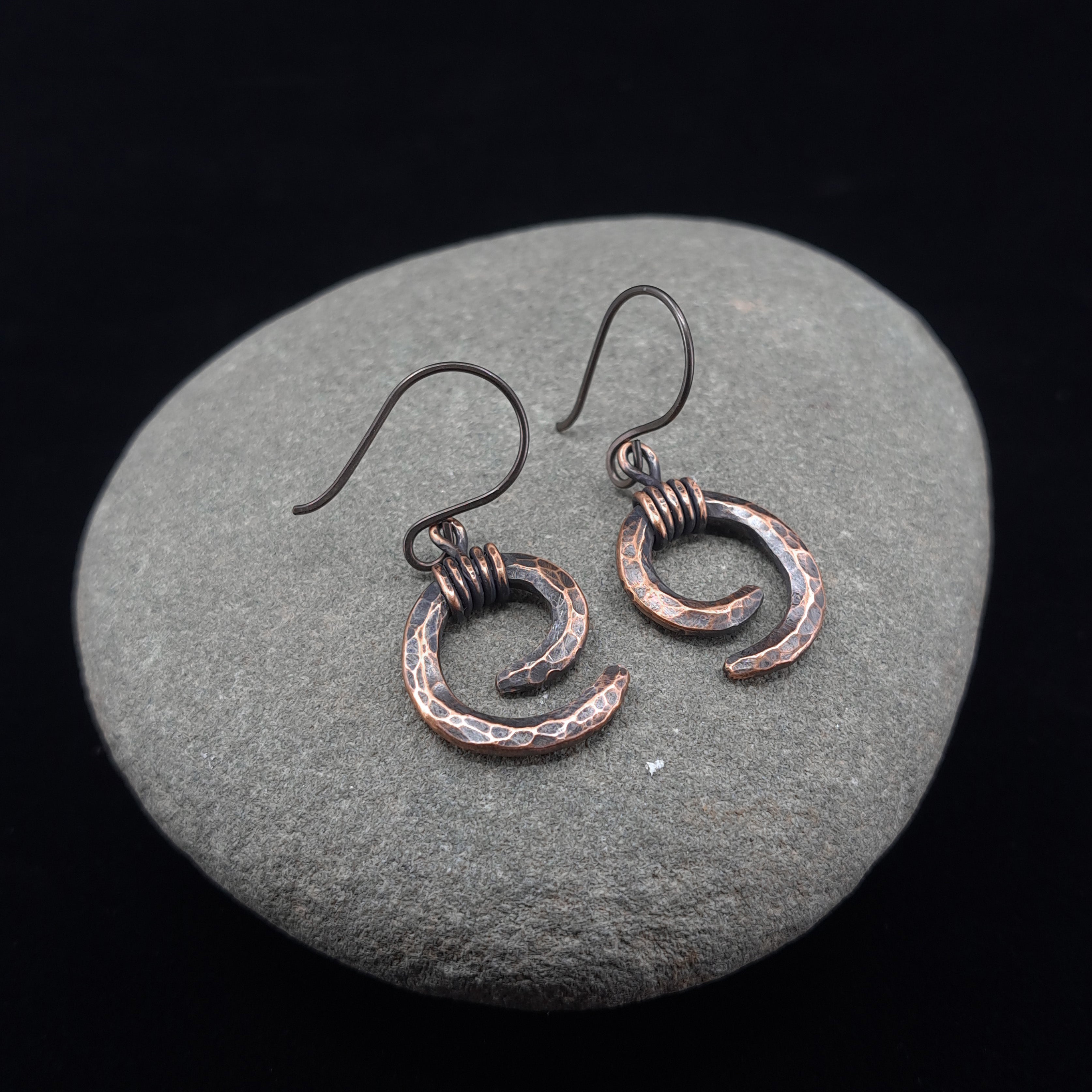 Hammered copper spiral earrings on river stone, dark background — Vortex