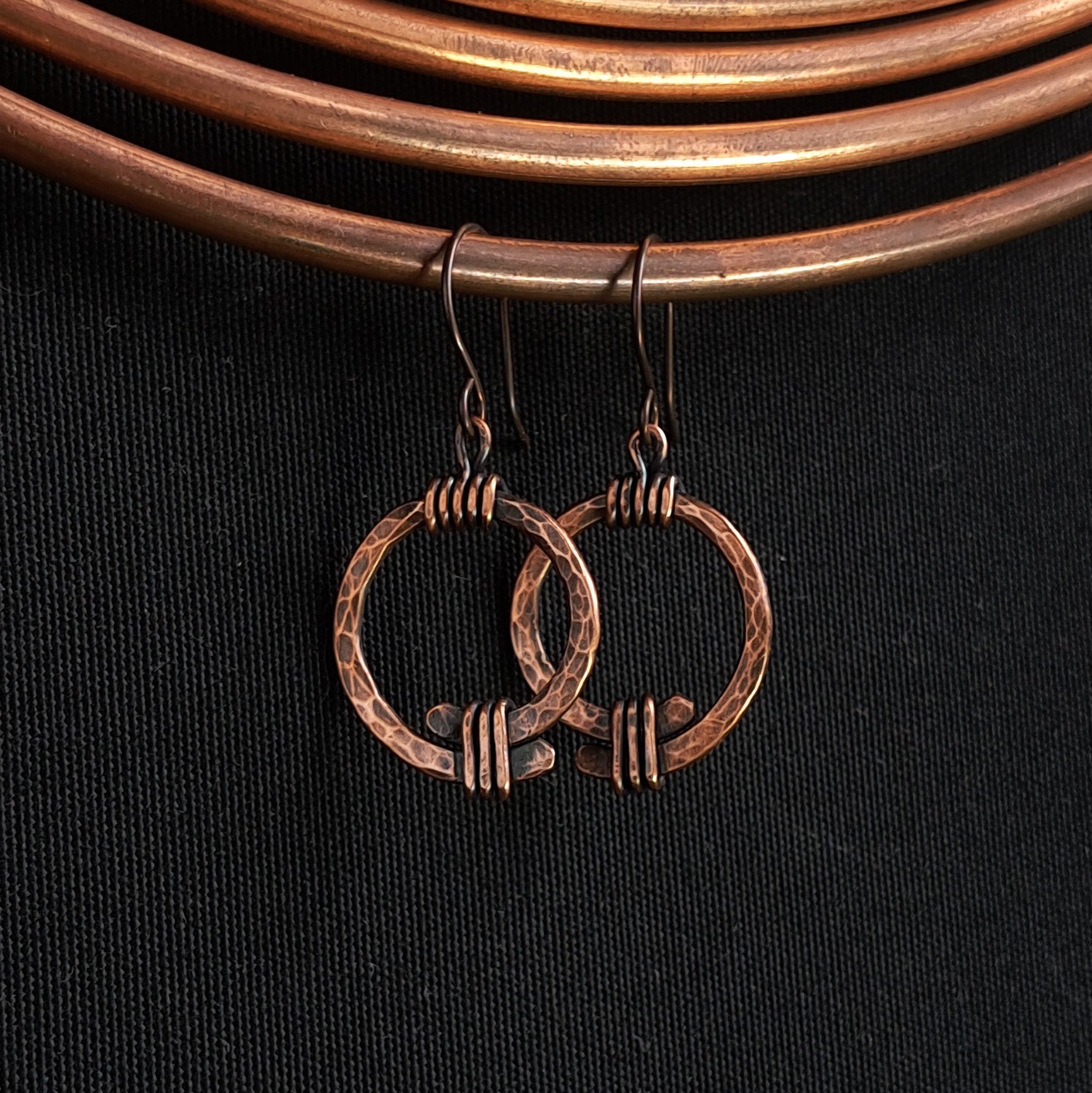 Copper open ring earrings hanging on copper coil, black fabric background — Torque