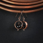 Hammered copper ring earrings with black onyx sphere on copper coil, dark fabric background — Meridian