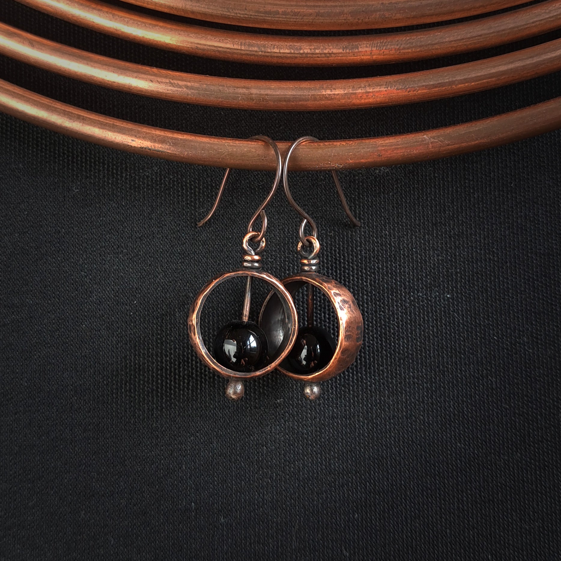 Hammered copper ring earrings with black onyx sphere on copper coil, dark fabric background — Meridian