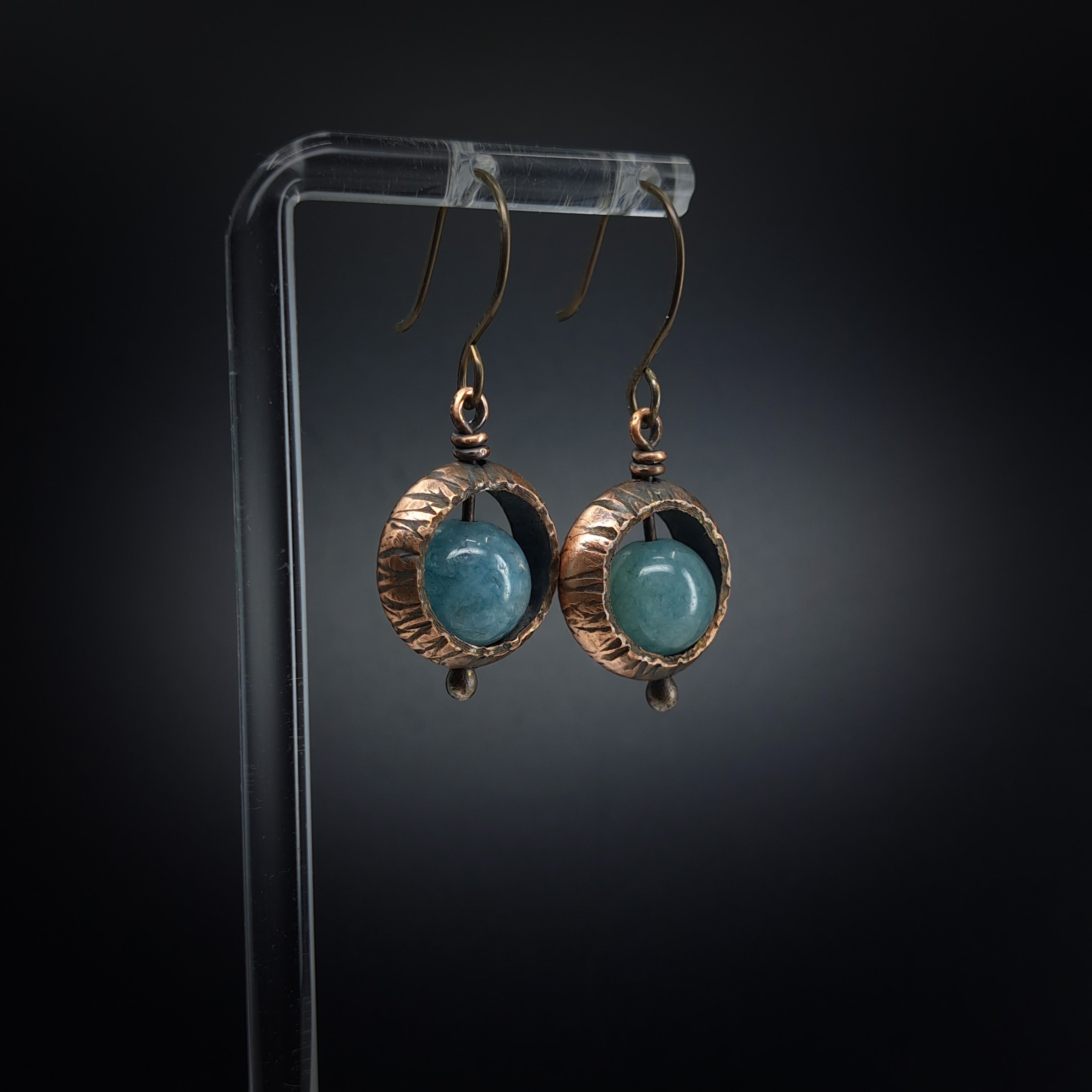 Copper ring earrings with aquamarine chalcedony on acrylic stand, dark background — side view, niobium hooks