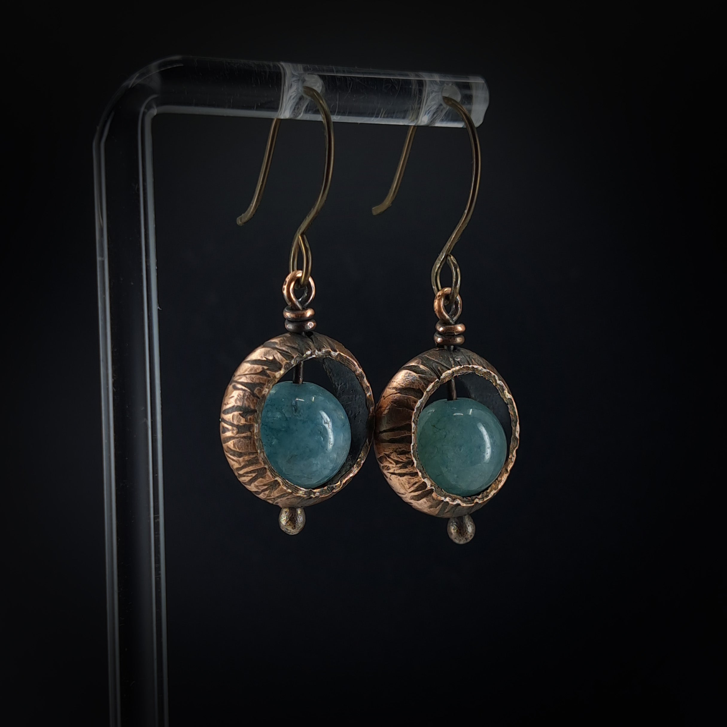 Copper ring earrings with aquamarine chalcedony on acrylic stand, black background — front view, oxidized copper