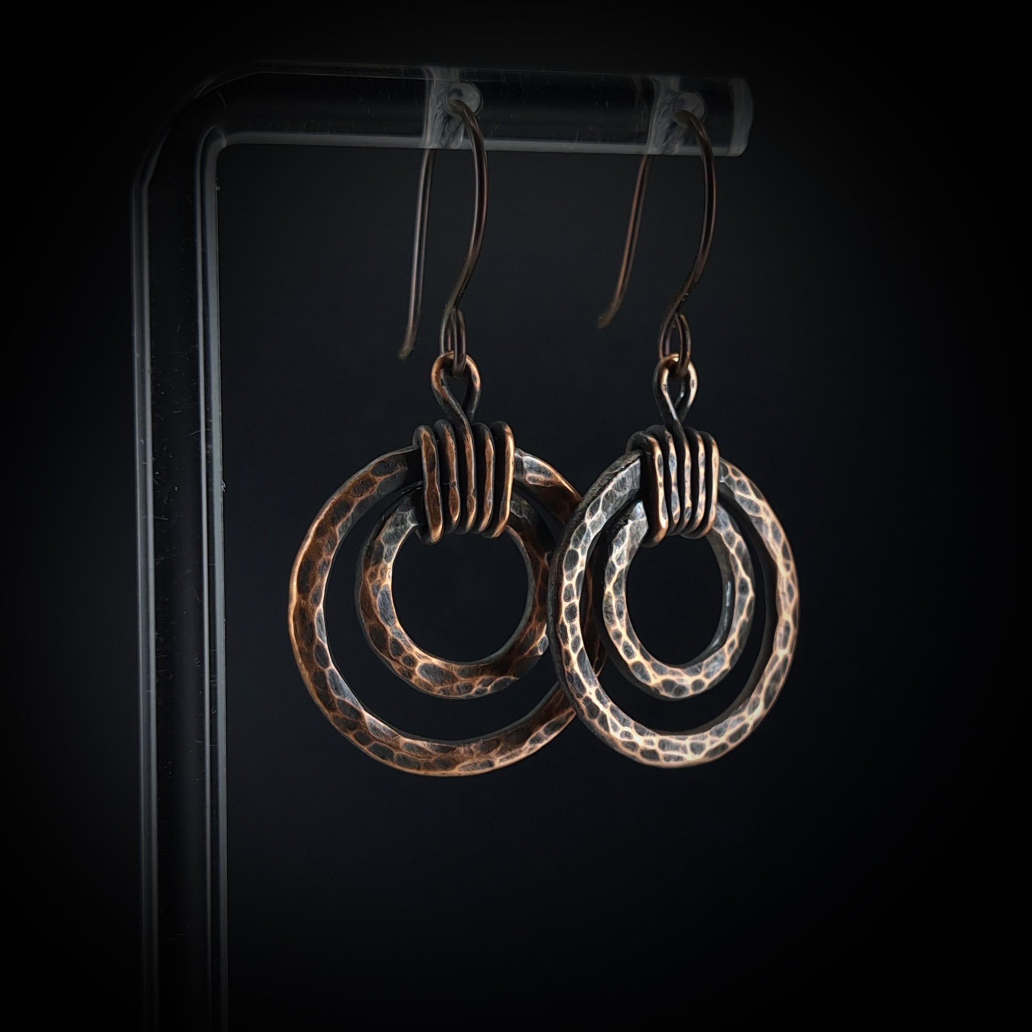 Hammered copper double hoop earrings on acrylic stand — dark background