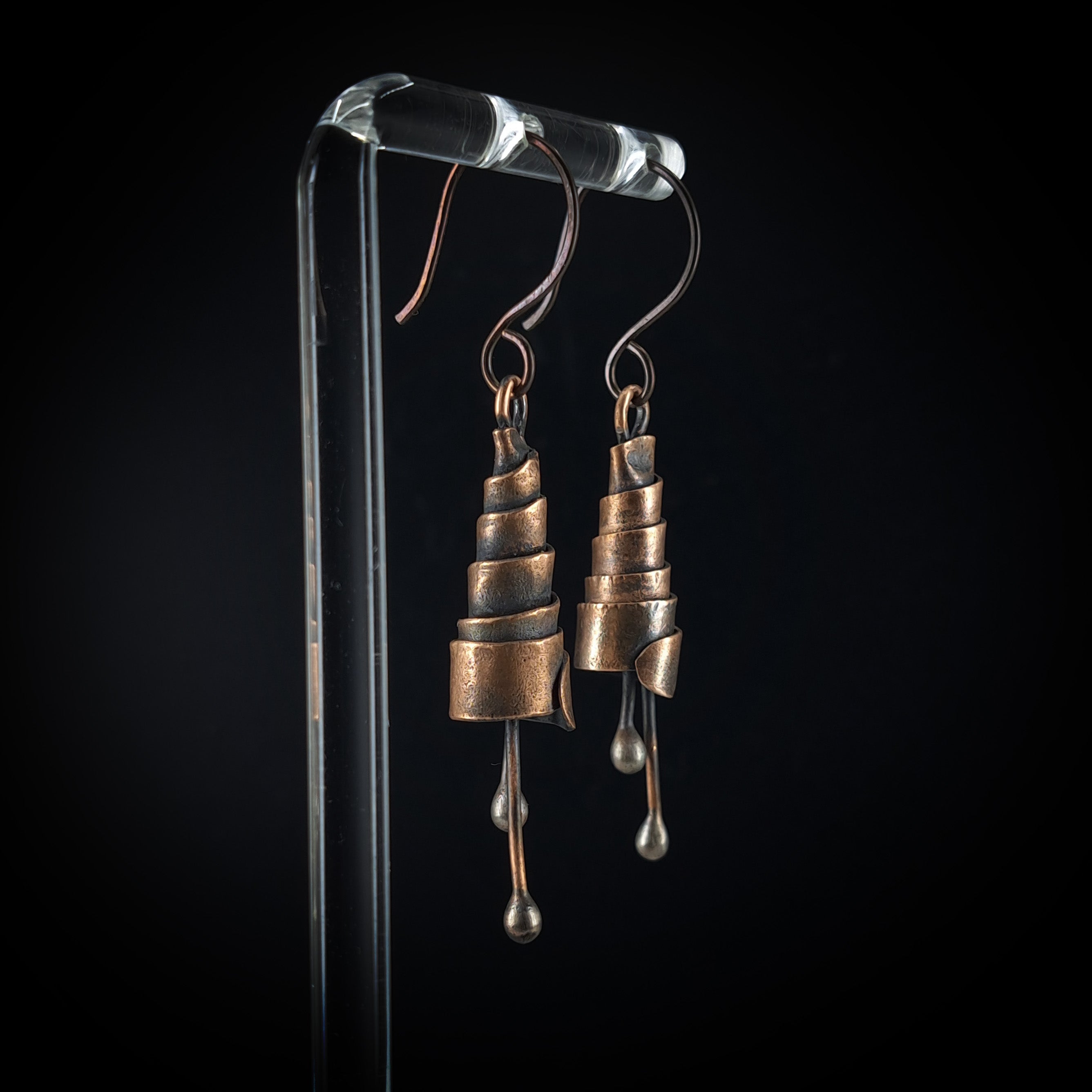 Copper spiral earrings on acrylic stand, black background — side view