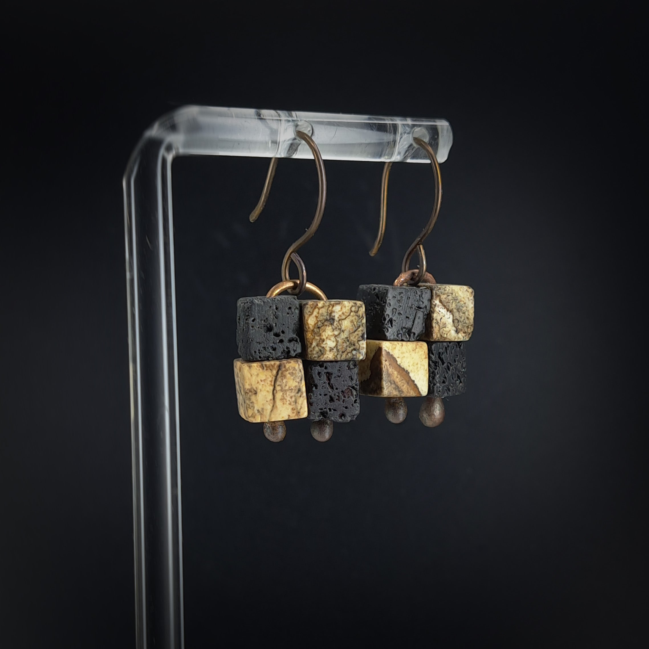 Lava stone and jasper earrings on white acrylic stand — side view, dark background
